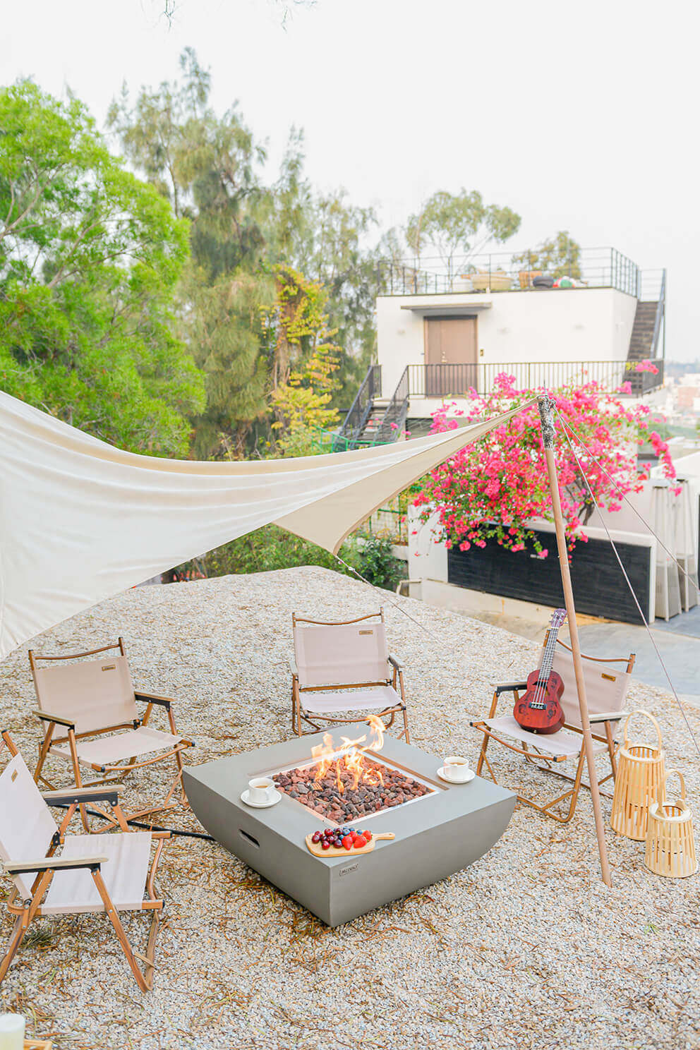 A light grey, square Modeno Westport Fire Table displayed outdoors on pale gravel, with a smooth glass-fibre reinforced concrete body and gently curved lower base. The central square burner tray is filled with reddish and charcoal-coloured lava rocks, with bright orange flames rising from the centre. Surrounding the table are beige folding camping chairs with wooden frames under a large cream-coloured shade sail, with a pink flowering bush and urban buildings in the background. The side of the fire table shows an inset hand-hold opening, emphasising its rounded lower form and modern silhouette.