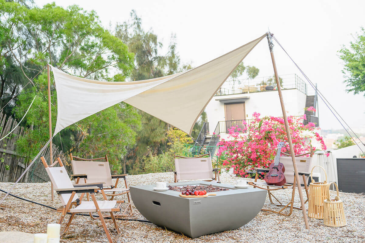 The Modeno Westport Fire Table positioned in an outdoor seating area beneath a large triangular shade canopy, surrounded by beige folding chairs with wooden frames. The fire table’s smooth, light grey body has curved sides and a recessed hand-hold on the visible face. The square stainless steel burner tray is filled with porous lava rocks, with a medium-height flame rising at the centre. Bright pink flowers and leafy trees frame the background, creating a warm, relaxed atmosphere.