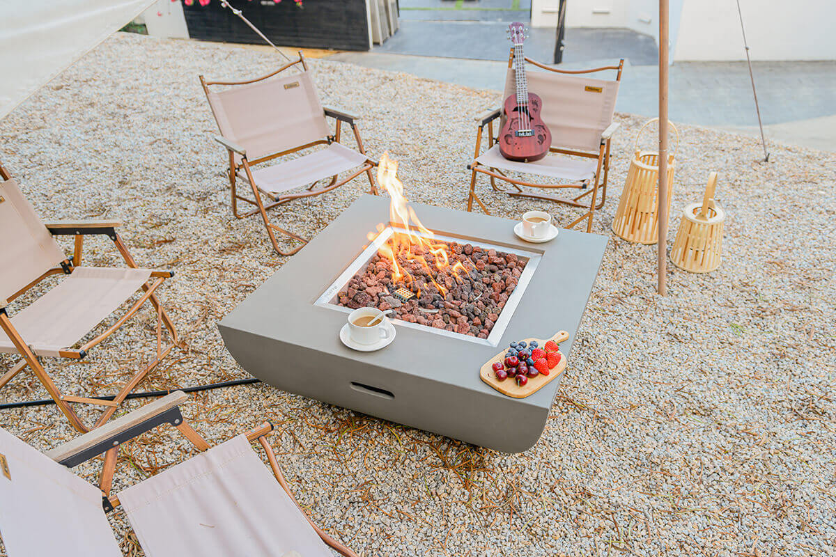 A high-angle view of the Modeno Westport Fire Table showing the entire square surface, with neatly arranged beige camping chairs forming a circle around it. The smooth light grey tabletop holds a central square burner surrounded by lava rocks, producing a twisting orange flame. A ukulele, wooden lanterns, and a gravel surface add natural textures to the setting. The table’s side hand-hold is visible, cut into the curved lower body for ease of movement.