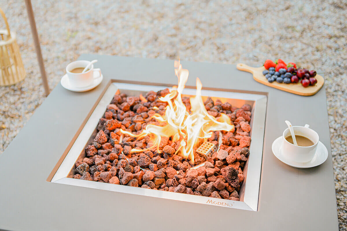 Close view of the square burner opening of the Westport Fire Table, showing lava rocks in varying red, brown, and charcoal tones arranged around the stainless steel burner tray. A bright, swirling flame rises from the centre. On the tabletop are two white coffee cups with matching saucers on opposite corners. A small wooden board holding strawberries, blueberries, grapes, and cherries sits along the front edge, contrasting against the light grey concrete surface.