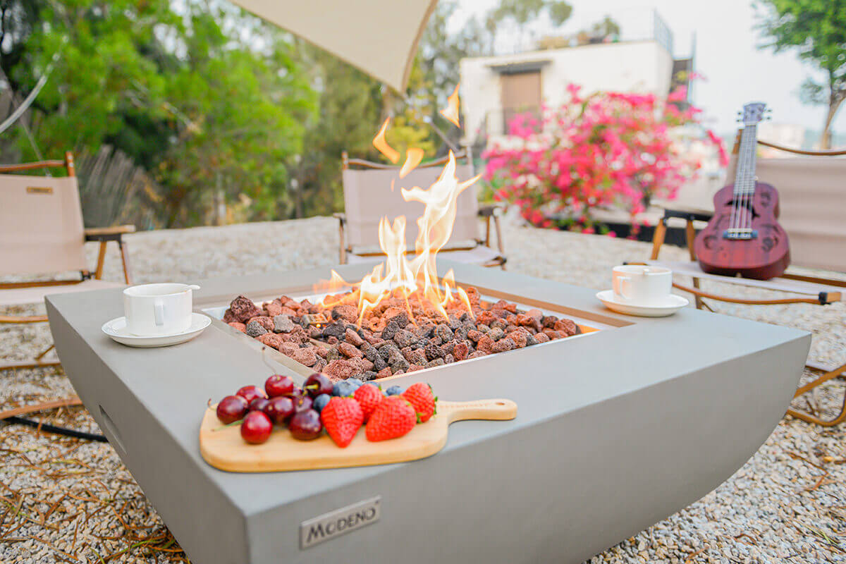 A low-angle closeup of the Modeno Westport Fire Table showing the smooth light grey concrete rim and the large square burner cavity filled with lava rocks. The central flame rises in layers of yellow and orange, with coffee cups and a board of fresh fruit arranged neatly on the tabletop. In the background, folding chairs and a ukulele create a relaxed outdoor gathering scene.