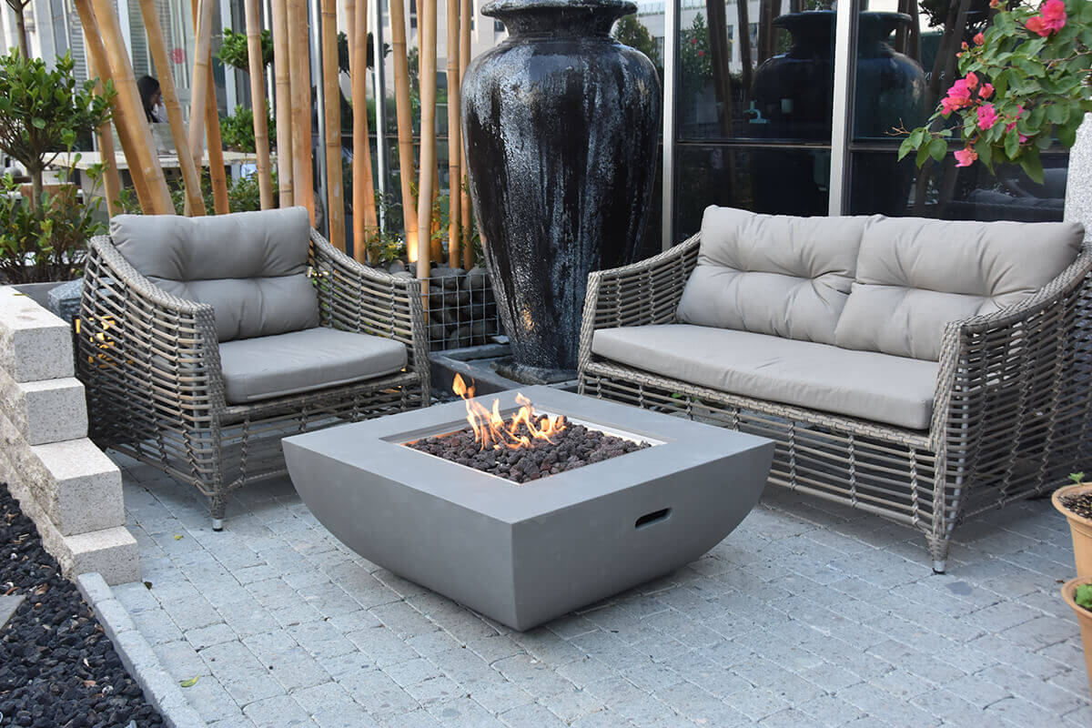 The Westport Fire Table placed in a contemporary outdoor patio with grey woven lounge chairs and a matching loveseat. The table’s curved concrete base and square lava rock-filled burner contrast with the clean stone paving. Tall bamboo stalks and a large dark ceramic water feature sit behind the seating area, adding depth and texture to the space while the medium-height flame burns steadily.