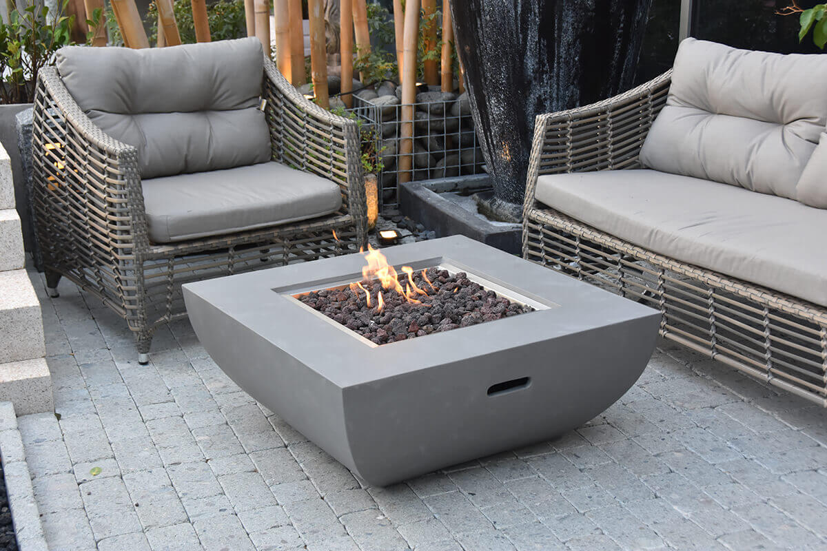 Another angle of the Modeno Westport Fire Table on a patio with grey woven outdoor seating. This view highlights the light grey, smooth concrete body with its gently curved lower half and the rectangular inset hand-hold along the side. The burner tray filled with dark, reddish lava rocks emits a warm, bright flame. The surrounding patio features grey brick pavers, bamboo, and a tall black ceramic vase, creating a spa-like environment.