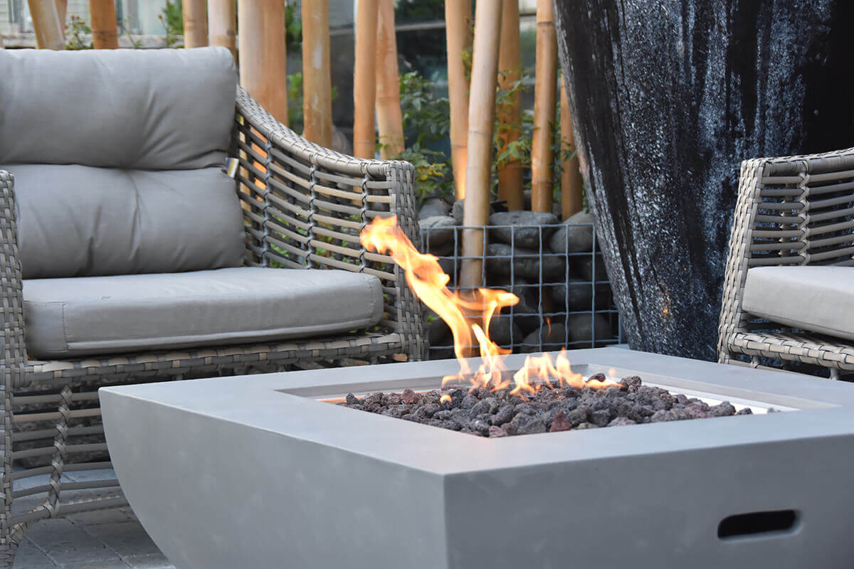 A closeup angled shot of the flame and lava rocks inside the Westport Fire Table’s square stainless steel burner tray. The smooth grey tabletop is visible in the foreground, with mid-height flames curving upward. Behind the fire table, woven grey outdoor chairs and tall natural bamboo poles create a calm, modern outdoor backdrop.
