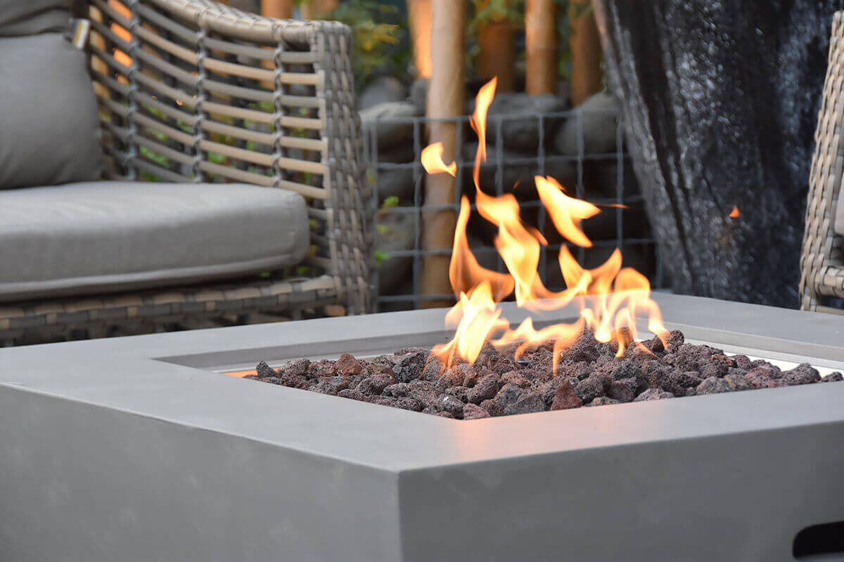 A tight closeup of the Westport Fire Table focusing on the flame rising from among rough-textured lava rocks. The shot captures the smooth light grey concrete rim of the table and the warm glow reflecting off the charred stones. Chairs and bamboo décor are softly blurred in the background, reinforcing the outdoor patio setting.