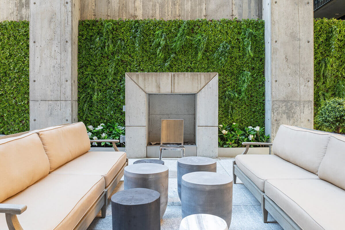 A close view of the Boxwood Privacy Trellis installed outdoors against a tall concrete wall, where dense artificial boxwood leaves in vivid medium and light green tones spread evenly across the entire panel. The trellis creates a full green backdrop behind modern patio seating that includes beige cushioned sofas and round stone like stools placed in the foreground. Soft natural light accentuates the layered leaf texture while small white flowers planted along the base soften the transition between the trellis and surrounding concrete architecture.