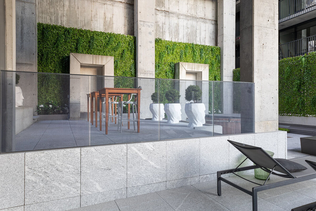 An outdoor architectural space featuring two large Boxwood Privacy Trellis panels installed vertically between tall concrete pillars. The dense artificial foliage forms vibrant green rectangles behind a raised patio area bordered by a glass railing. A tall wooden table with clear modern stools sits on the terrace, accompanied by sculptural white planters holding small manicured topiary shrubs. The scene shows how the trellis provides both privacy and decorative contrast against the raw concrete surfaces.