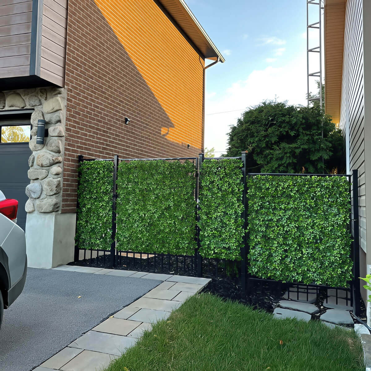 A residential side yard where three Boxwood Privacy Trellis panels are mounted along a black metal frame fence beside a brick home. Each panel forms a dense wall of small, glossy green artificial leaves arranged tightly to block the view between properties. The panels sit above dark mulch and stone edging, with the driveway and lawn visible in the foreground. The realistic texture of the foliage provides an immediate privacy solution for compact suburban spaces.