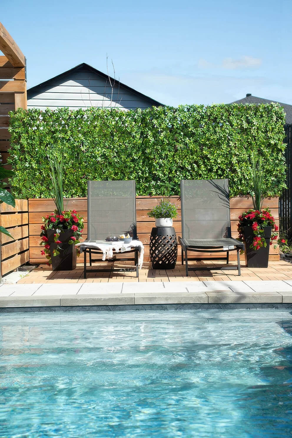 A bright poolside scene where a long Boxwood Privacy Trellis installation creates a tall hedge like backdrop behind two grey mesh loungers. The dense artificial boxwood foliage stretches across the entire width of the sitting area, positioned on a wooden deck above the pool. Large black planters filled with red flowers and tall green leaves frame both sides of the loungers, with the vivid greenery of the trellis providing a natural looking enclosure for the sunbathing space.
