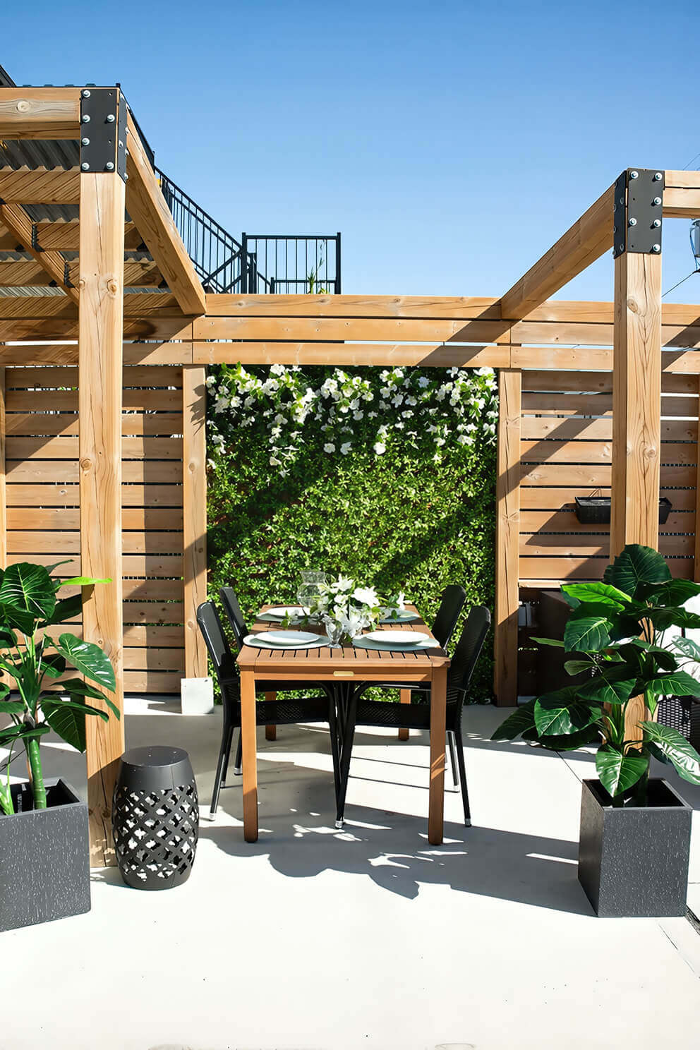 A patio dining area beneath a wooden pergola, where a Boxwood Privacy Trellis forms a full vertical green wall behind a small wooden dining table set for four. The artificial foliage consists of tightly packed small leaves in shades of medium and light green, with clusters of white flowers draping over the top portion of the trellis. The trellis adds a lush garden effect to the structured wooden surroundings while maintaining privacy for the seating area.