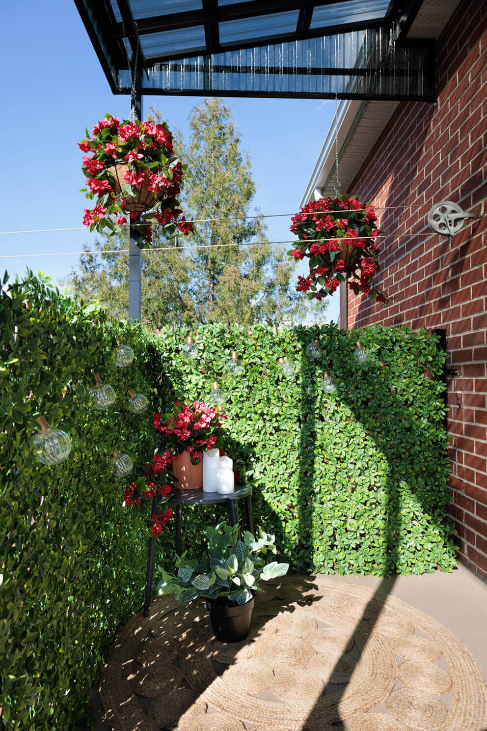 A compact balcony lined with Boxwood Privacy Trellis panels arranged in an L shaped corner configuration. The artificial leaves cover the entire railing height to create privacy from surrounding properties. Warm sun casts shadows over the foliage while decorative string lights hang across the trellis. Two hanging baskets filled with bright red flowers frame the top of the scene, and a small side table displays potted plants and candles, enhancing the enclosed garden ambience.