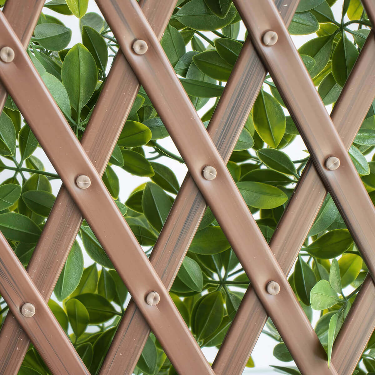 A zoomed in view of the Boxwood Privacy Trellis showing the brown plastic lattice arranged in a repeating criss cross pattern, with each intersection secured by small rounded fasteners. Behind the lattice, densely packed artificial leaves in multiple green tones push through the openings, creating a natural appearance while revealing the structure that allows the trellis to expand and contract.