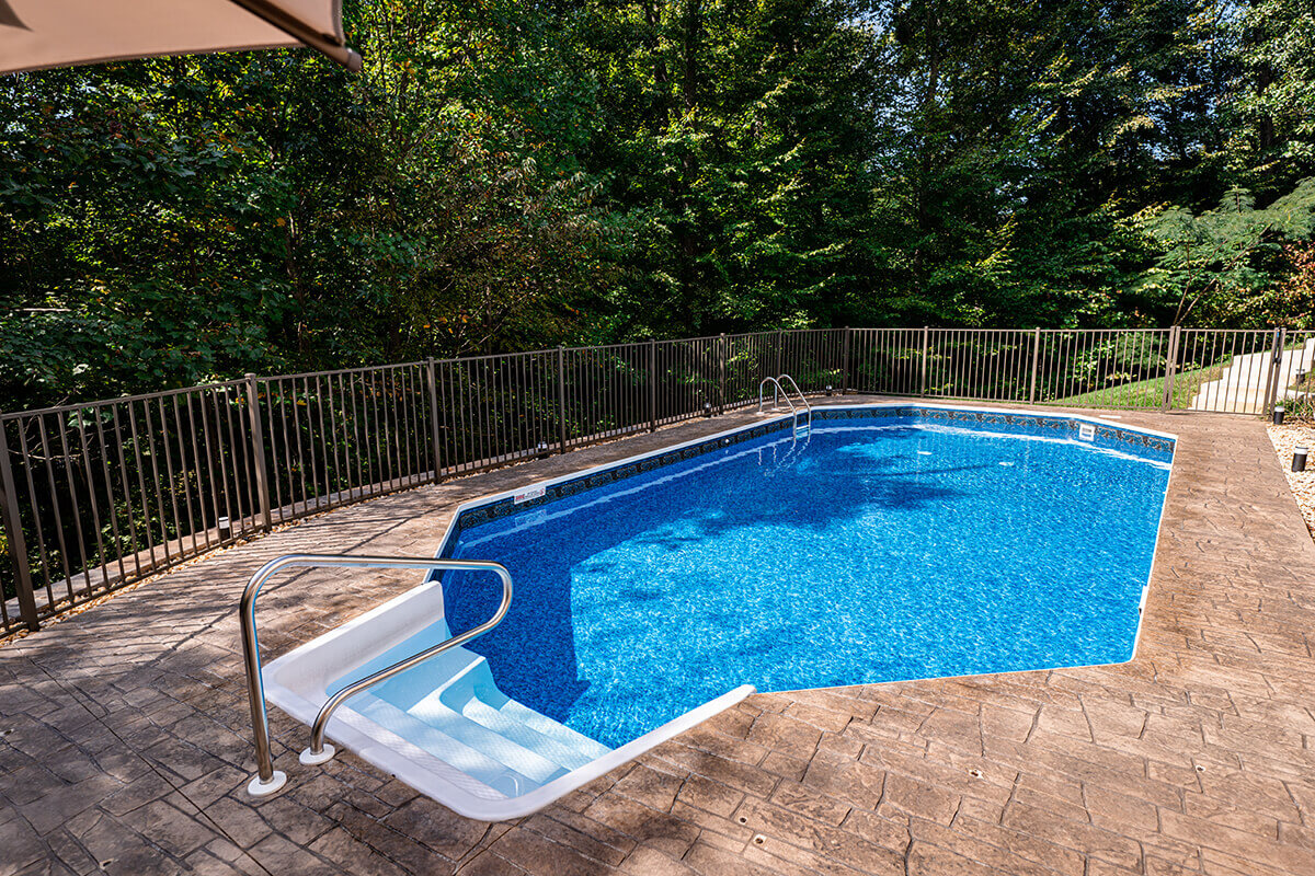 Fox Ultimate Pool installed as an inground Grecian-shaped pool surrounded by textured stamped concrete decking, with white thermoplastic entry steps located at the shallow end on the left side, featuring a stainless steel handrail installed into the deck. The pool water is bright blue with a patterned vinyl liner visible at the waterline. A continuous metal safety fence runs along the perimeter of the pool area, backed by dense green trees creating natural privacy.