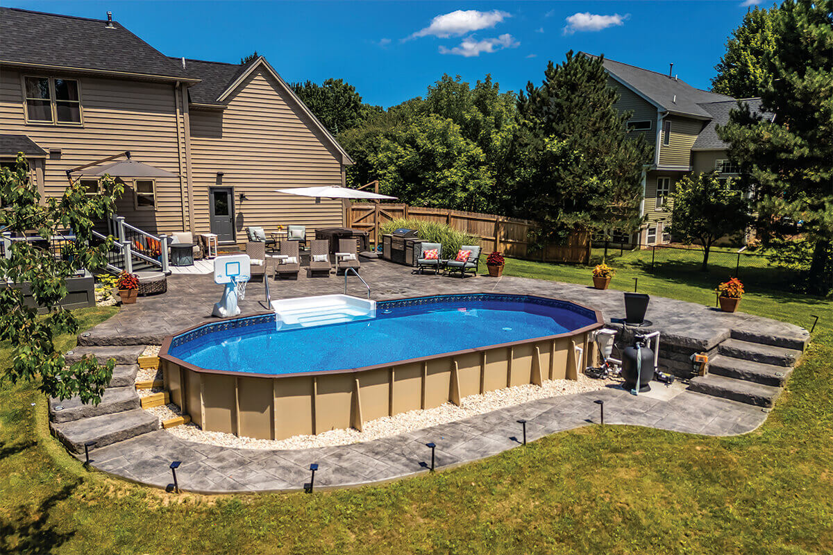 Fox Ultimate Pool installed semi-inground in an oval shape with exposed tan steel wall panels and structural buttress supports visible along the sides. The raised installation is bordered by a curved stamped concrete patio with outdoor furniture and a basketball hoop near the shallow end. A white thermoplastic step system is installed at the front of the pool, leading into clear blue water. The backyard features a large lawn, potted plants, and a deck attached to a beige house in the background.