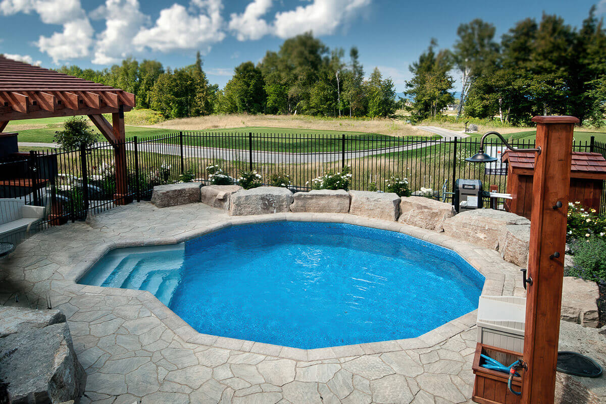 Fox Ultimate Pool installed inground in a Grecian shape, surrounded by natural flagstone decking and large boulder accents along the far side. White thermoplastic steps with wide treads are positioned in the near corner, descending into the clear blue water. A black metal fence encloses the pool area, with open green landscape and a tree-lined horizon extending beyond the fenced yard.