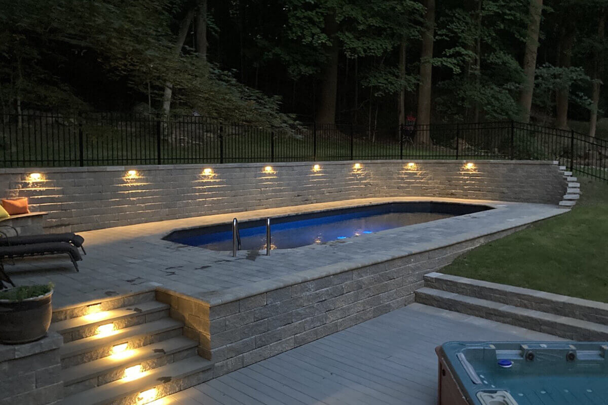 Fox Ultimate Pool installed semi-inground with a raised stone retaining wall on one side, creating a recessed pool area illuminated by multiple warm deck lights. The oval-shaped pool features dark blue water reflecting the evening sky. A metal handrail leads into the pool at the shallow end. Surrounding landscaping includes trees, seating areas, and outdoor steps integrated into the stonework.