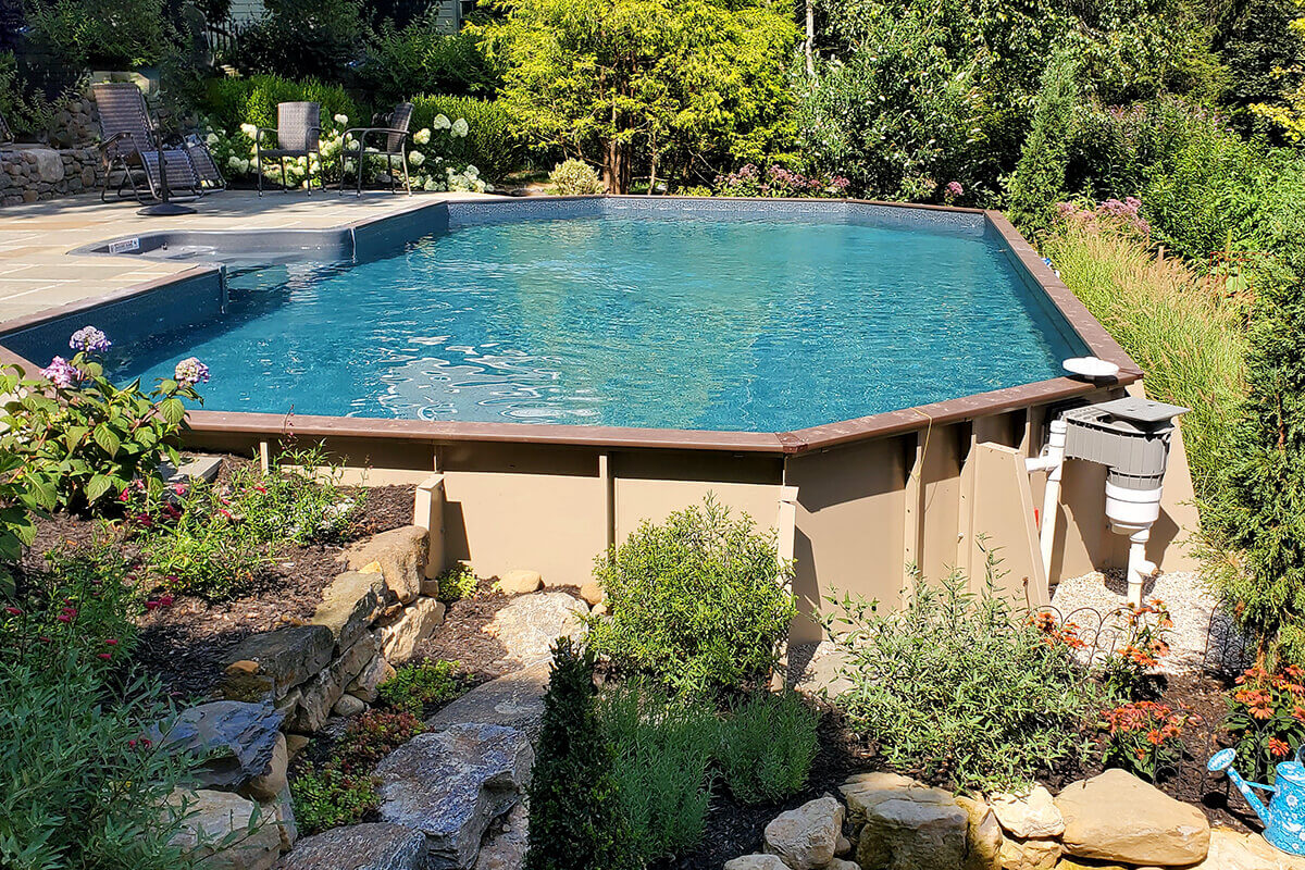 Fox Ultimate Pool installed semi-inground with tan steel wall panels and structural supports fully visible on the exterior. The water appears deep blue, with landscaping featuring shrubs, flowers, and stacked stone borders surrounding the pool area. Adjacent patio furniture sits on flat stone pavers, and equipment is visible near the pool wall on the right side.