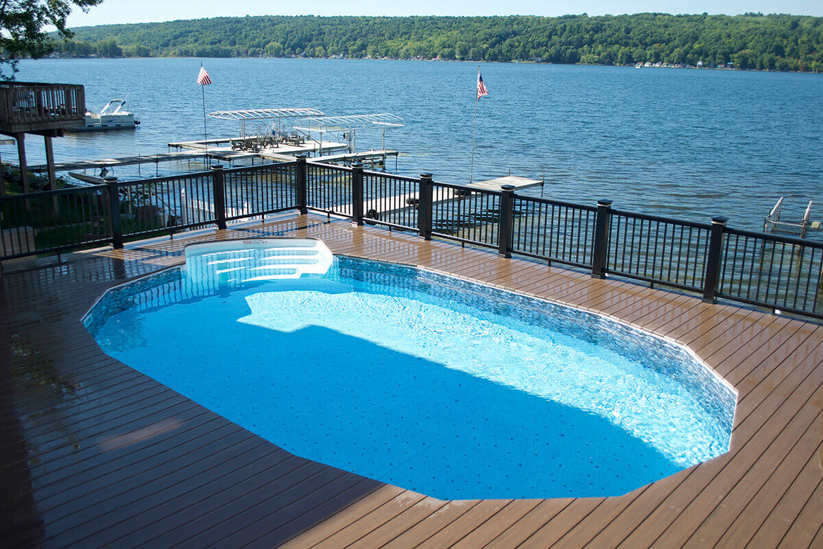 Fox Ultimate Pool installed inground on a raised wooden deck overlooking a lake. The oval pool features light blue water with a patterned vinyl liner visible along the top edge. White thermoplastic entry steps extend from the near side into the shallow end. Black railings run along the deck perimeter with docks, boats, and tree-covered hills in the background.