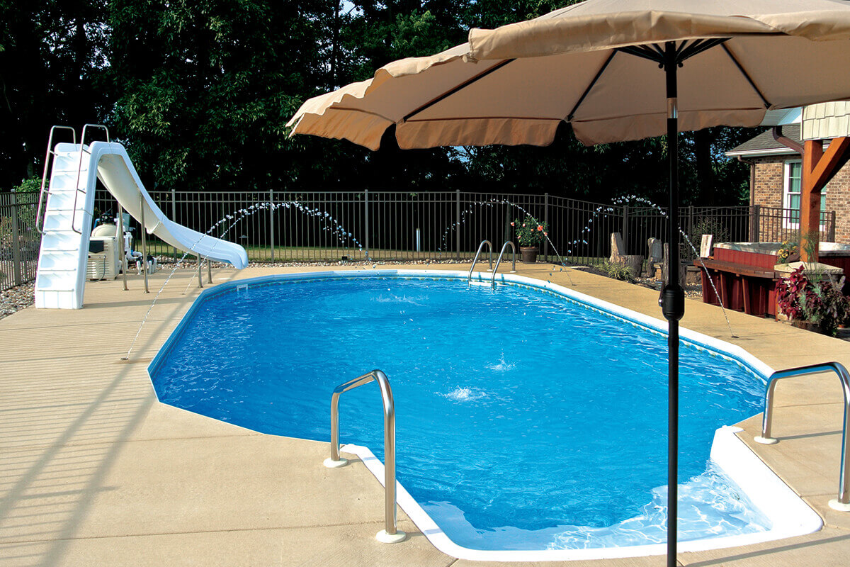 Fox Ultimate Pool installed inground in an elongated oval shape, surrounded by a beige concrete patio. A large beige umbrella shades part of the deck on the right, while a tall white slide at the far end leads into the pool. Multiple arched water features spray streams across the pool surface. Stainless steel handrails are installed at both ends for safe entry.