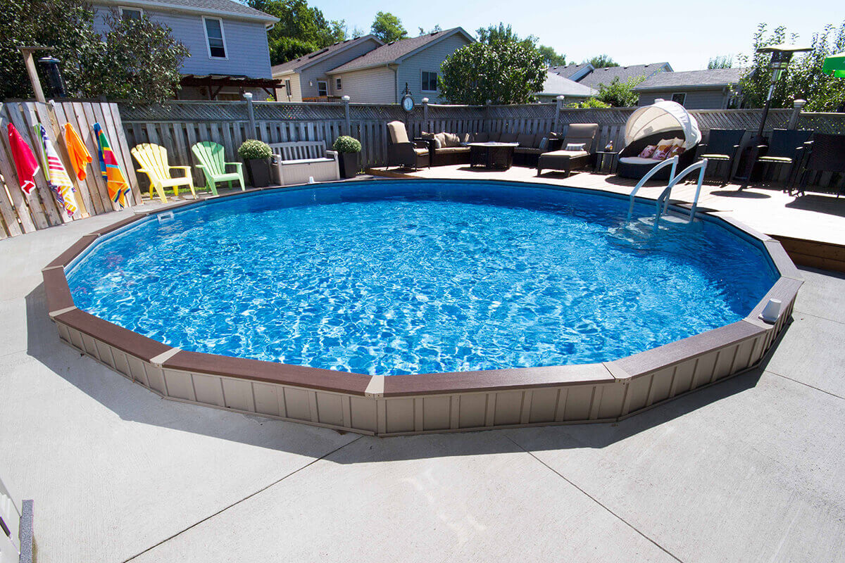 Fox Ultimate Pool installed above ground in a round shape, featuring tan steel wall panels and brown top rails framing the bright blue water. The surrounding patio includes concrete slabs and a wooden deck area with outdoor seating. A metal ladder provides access at the far side, and colourful towels hang from a wooden privacy fence in the background.