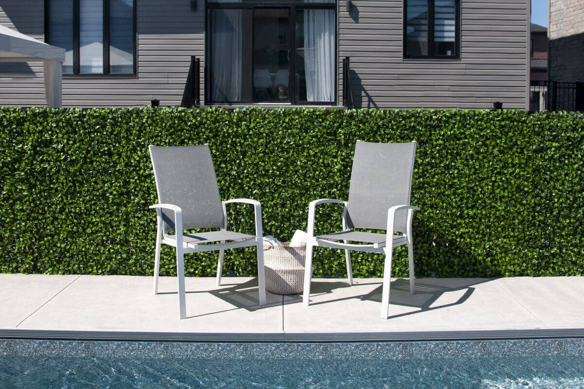 A pair of tall outdoor panel screens covered in dense artificial green foliage arranged along the edge of a residential poolside patio. The panels are mounted against a low concrete border wall, providing a uniform green backdrop behind two grey mesh outdoor chairs positioned on a concrete surface. The pool water in the foreground reflects sunlight, and a modern house exterior sits behind the foliage. The faux leaves are tightly layered, creating opaque privacy coverage with varying green tones.