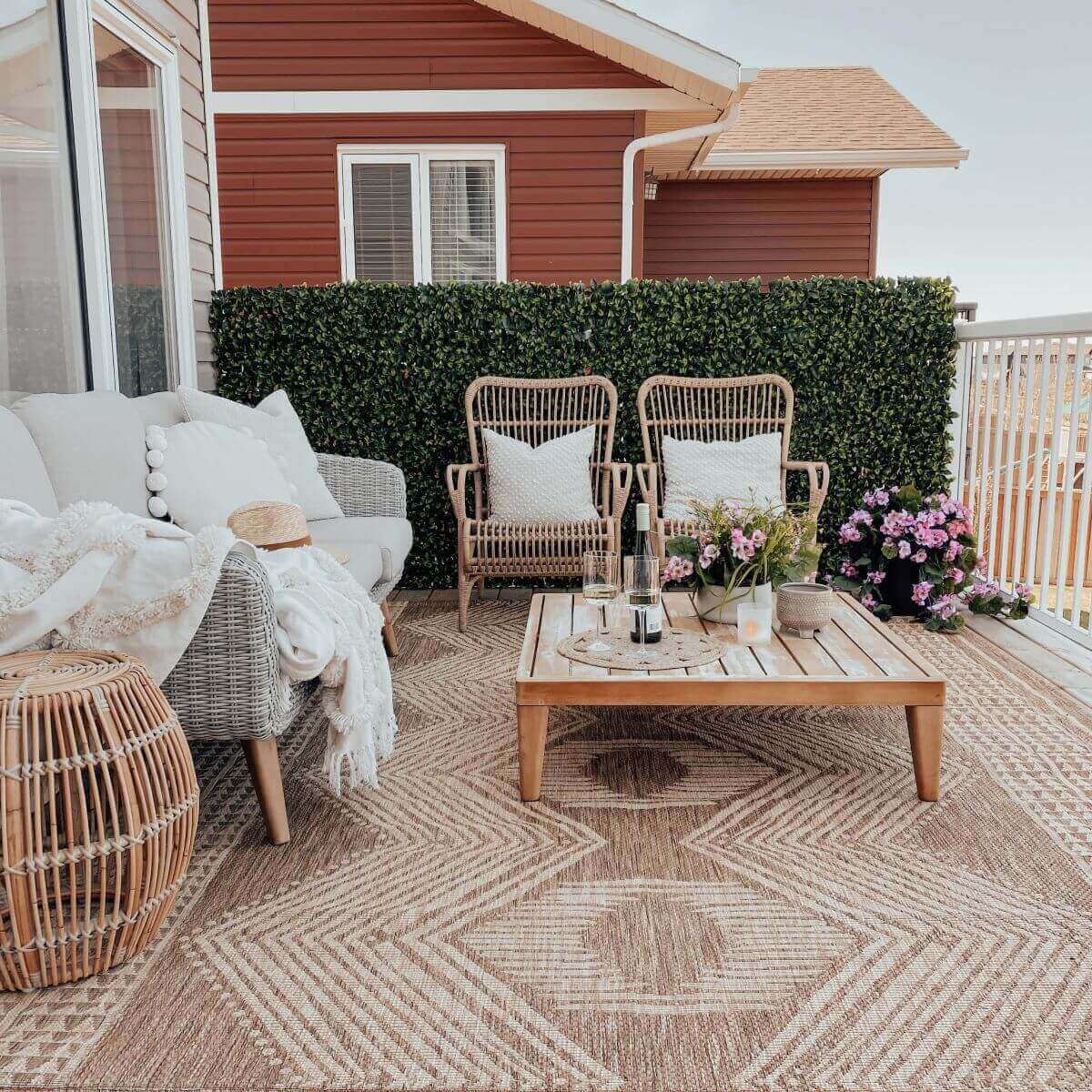 A cosy balcony seating area enclosed by a long stretch of artificial green privacy panels attached along the railing. Two natural wicker armchairs sit side by side with white cushions, facing a low wooden coffee table decorated with drinks, flowers, and a tray. The scene includes a soft rug under the furniture and a red house exterior behind the panels. The foliage panels feature tightly packed faux leaves with subtle colour variations that create a natural and consistent green wall.