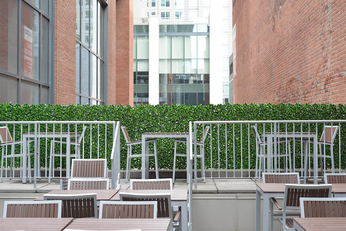 A commercial patio space bordered by a continuous row of artificial green privacy panels mounted along a raised platform with metal railings. Multiple outdoor dining tables with brown slatted tops and grey metal frames fill the foreground, while matching tall café tables sit behind the railing. The dense faux foliage provides screening between tall brick buildings and a glass office structure in the background. The leaves display mixed green hues and are arranged densely for full visual coverage.