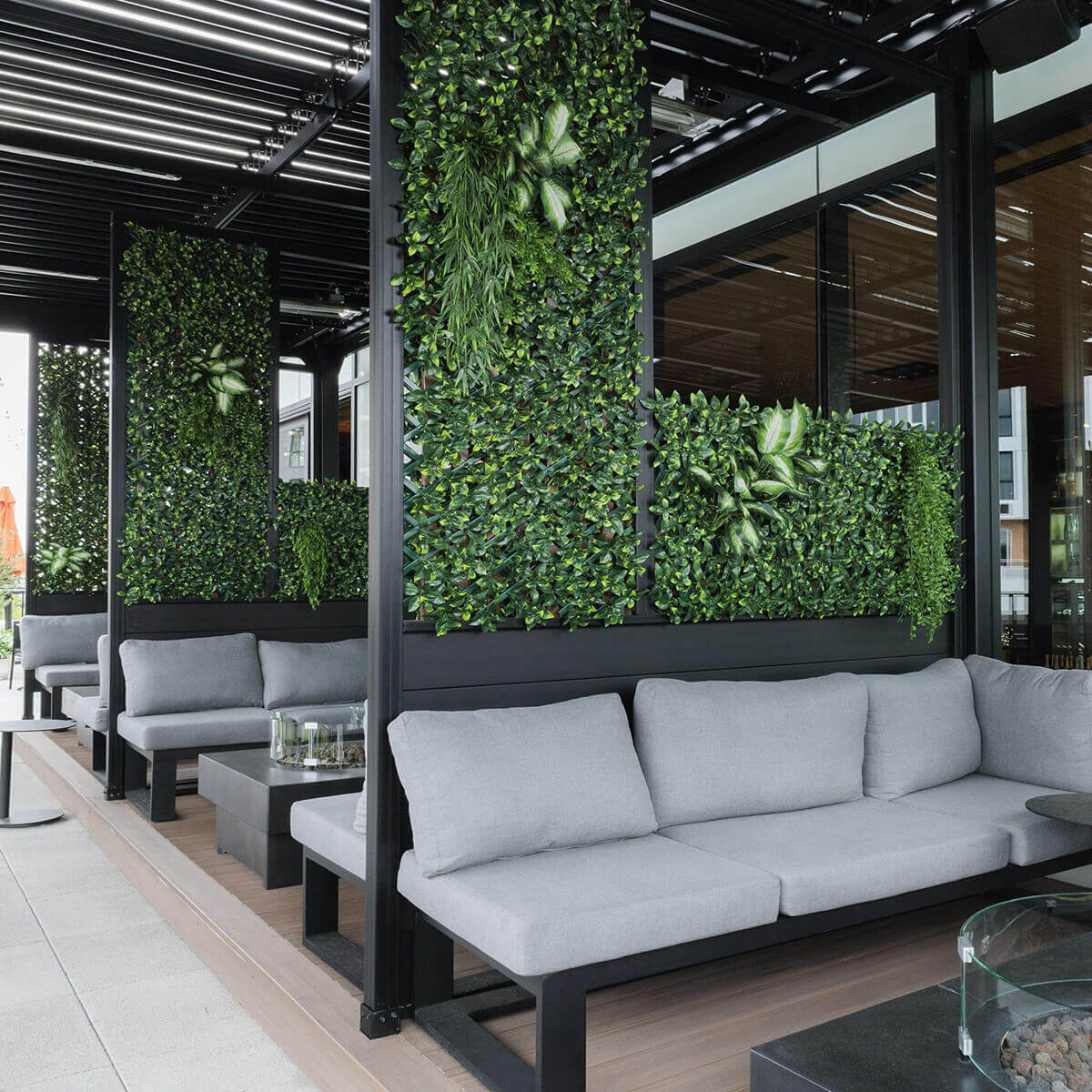 A stylish outdoor lounge featuring several vertical rectangular privacy panels installed within a modern black pergola structure. Each panel is filled with dense artificial green foliage, with some areas accented by integrated faux plants that add depth and texture. Grey cushioned sectional seating sits below the panels, creating a semi enclosed lounge space. The foliage varies in leaf size and shade, forming a layered green wall with strong visual density and decorative appeal.