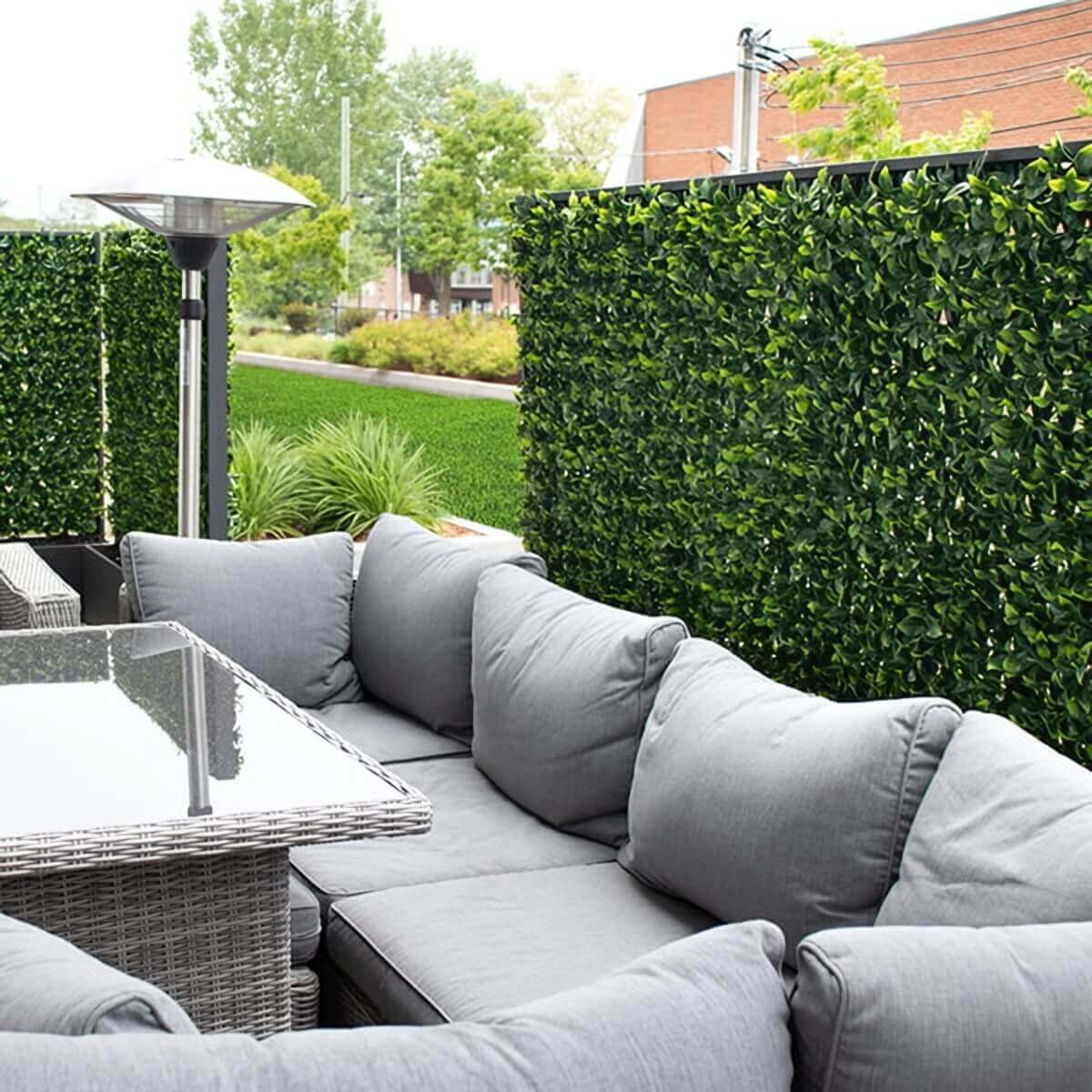 A contemporary patio seating area bordered by tall artificial green privacy panels placed behind a grey wicker sectional sofa with plush cushions. A glass topped dining table sits in front of the sofa, and a stainless steel patio heater stands near the left side. The faux foliage forms a continuous, opaque green barrier with closely packed leaves in bright and dark green tones, adding privacy against a backdrop of trees and rooftops.