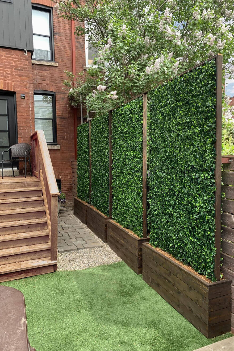 A narrow urban backyard lined with three tall privacy screens mounted inside raised wooden planter boxes. Each panel is densely filled with artificial green leaves, creating full height coverage along the boundary. The area features a wooden staircase leading to a back door, a gravel walkway, and a small patch of synthetic grass. The leaves vary slightly in shape and shade, forming a realistic and completely opaque green barrier that blends with surrounding trees.