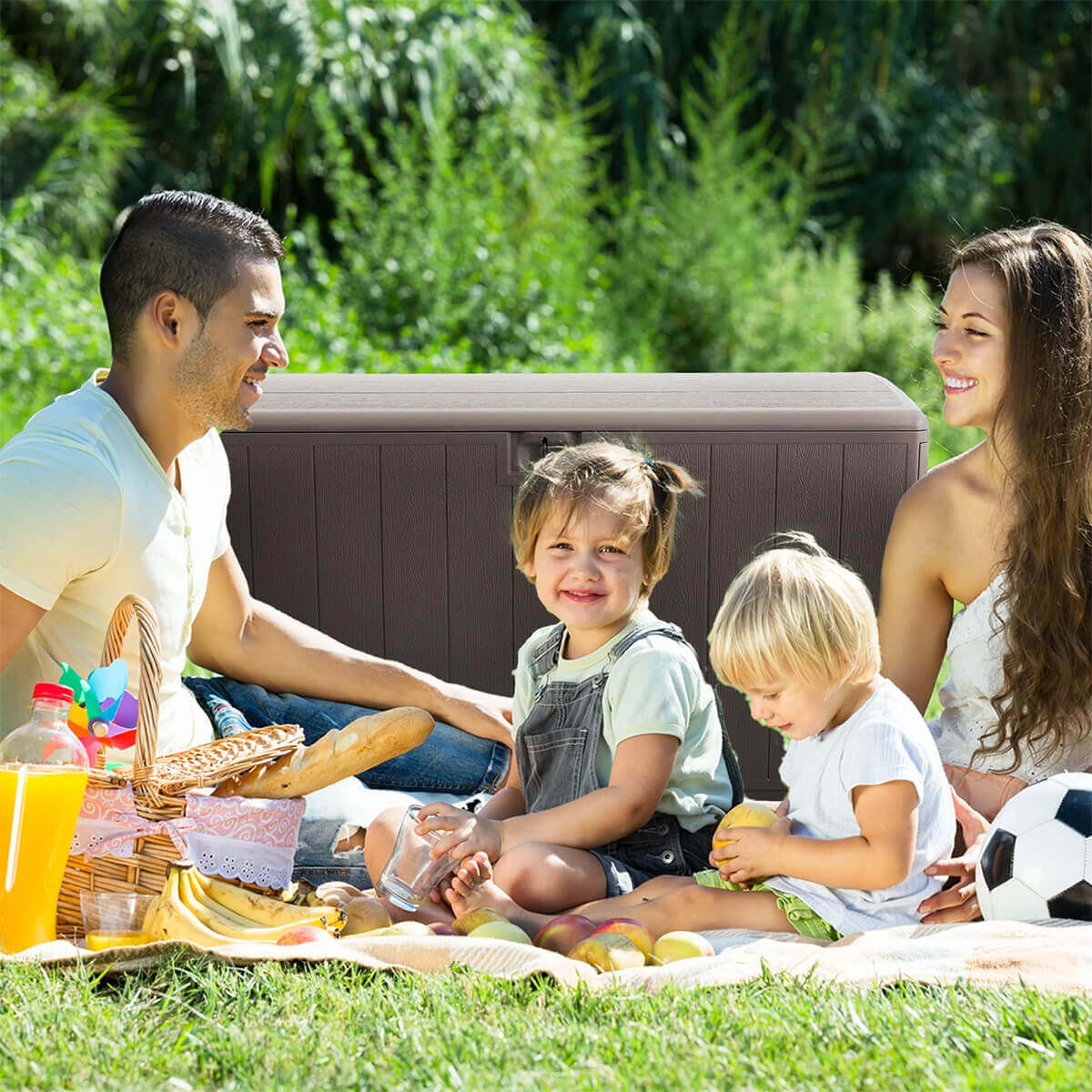 PDG 105 Gallon Resin Outdoor Storage Deck Box positioned behind a family enjoying a picnic on a grassy lawn. The driftwood-coloured deck box forms the backdrop, partially obscured by two young children seated on a blanket along with picnic food, fruit, and a football. Vertical panel detailing and the curved lid of the storage box are visible above the children’s heads.