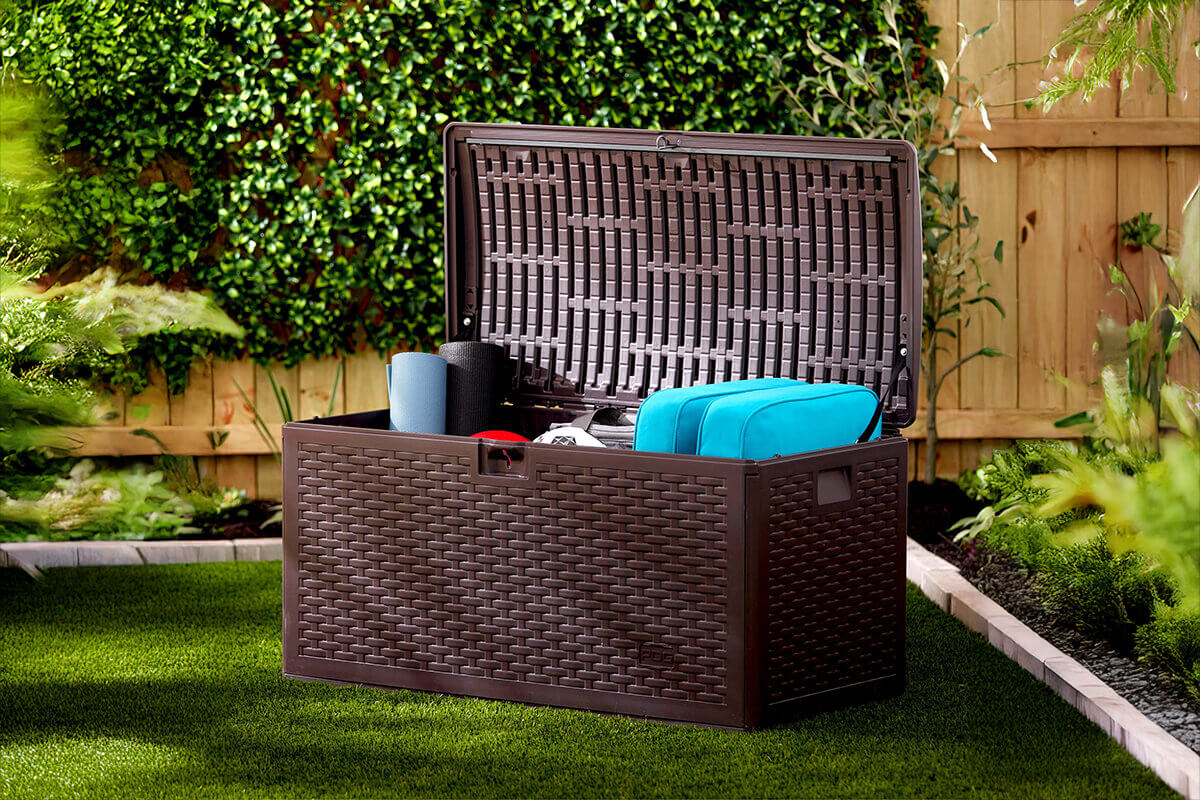 PDG 73 Gallon Resin Outdoor Storage Deck Box placed on a vibrant green lawn in a landscaped garden. The lid is raised fully, supported by the internal retainer straps. The interior holds several items including rolled exercise mats, bright turquoise outdoor cushions, and small accessories. The front and side panels feature the detailed wicker-pattern resin texture, and the moulded frame sits on trimmed edge pavers bordering the lawn.