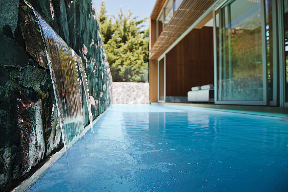 Image representing Pool Pumps in use, featuring a modern in ground swimming pool with a smooth sheet style waterfall flowing from a dark stacked stone wall on the left directly into clear blue pool water below. The continuous curtain of moving water creates visible surface ripples, illustrating active water circulation and consistent flow driven by pool pump operation. The pool stretches toward a contemporary home in the background, with large glass sliding doors and warm wood exterior panels on the right, reinforcing the role of pool pumps in maintaining clean, evenly circulated water within a high end residential pool setting.