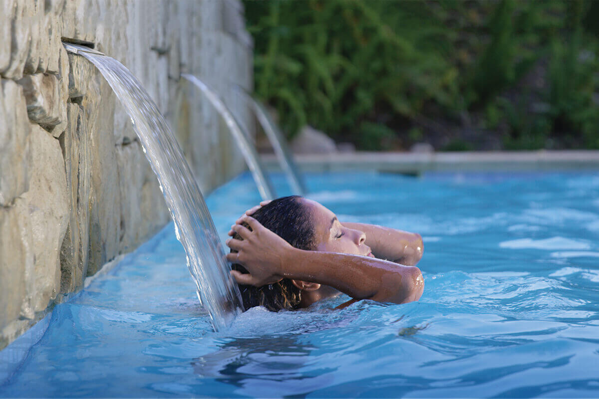 In-ground swimming pool scene illustrating the role of in-ground pool pumps, showing a built in stone wall with multiple narrow waterfall spillways pouring steady streams of water into clear blue pool water. A swimmer rests beneath one of the flowing water features, with water cascading over their upper back and shoulders, demonstrating strong circulation and continuous flow supported by in-ground pool pump operation. The evenly moving water surface and active waterfalls visually reinforce how in-ground pool pumps power consistent circulation, enhance water clarity, and support smooth day to day performance in a residential in ground pool environment.