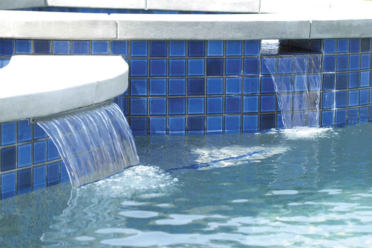 Commercial Pool Pumps supporting a high-use swimming pool, shown through multiple sheer-descent water features evenly spaced along a curved stone pool wall. Clear sheets of water flow continuously from horizontal spillway openings set just beneath the coping stones, creating smooth cascades into the pool below. The stone veneer wall wraps around the pool’s perimeter, with each waterfall aligned at equal height and distance, demonstrating consistent water pressure and balanced circulation typically maintained by commercial pool pump systems in professional aquatic environments.