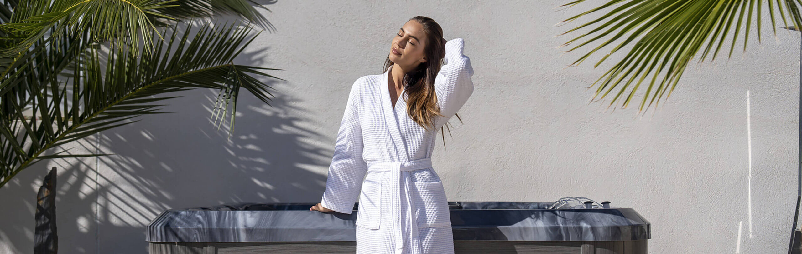 Lifestyle banner image of a woman standing beside a Maple Spas hot tub in a modern outdoor setting, captured in a wide horizontal format. The woman is positioned slightly right of centre, wearing a white textured spa robe tied at the waist, with her left hand resting on the edge of the hot tub and her right hand gently lifting her hair as she tilts her face toward the sunlight. She appears relaxed and content, eyes closed, conveying a sense of calm and rejuvenation. The hot tub features a dark marbled acrylic shell edge and grey horizontal wood-grain cabinet panels beneath. A waterfall feature is visible along the top right edge of the spa, with water softly cascading into the tub. The background consists of a smooth light-coloured exterior wall framed by large green palm leaves extending inward from both the left and right sides, casting natural shadows across the wall and spa surface. Bright daylight creates strong, clean highlights and defined shadows, reinforcing a premium outdoor wellness atmosphere associated with Maple Spas relaxation and luxury spa living.