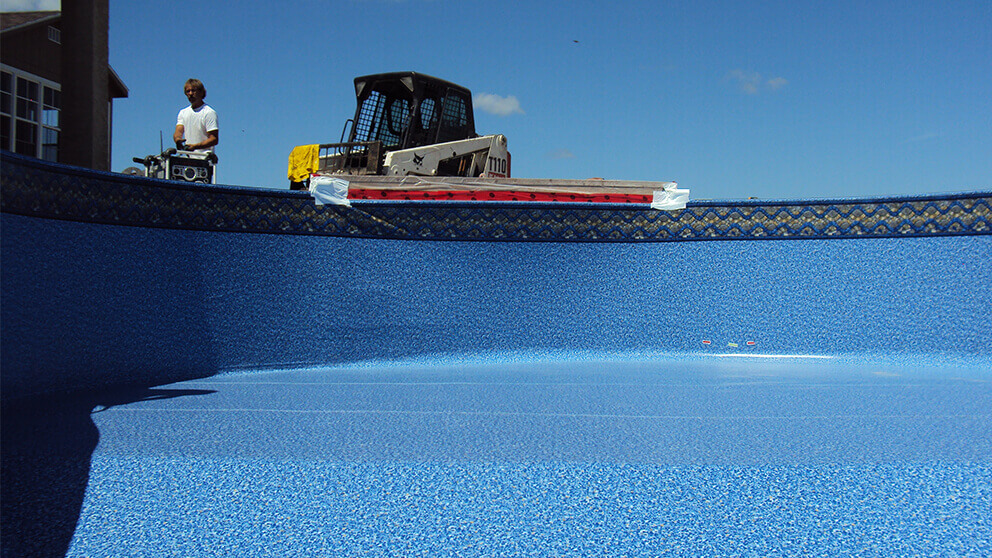 This high angle view from inside an empty in-ground swimming pool captures a professional Winnipeg pool liner replacement completed by Crown Spas and Pools. The interior surface is fully covered in a new bright blue vinyl liner with a fine granular texture that stretches across the curved walls and pool floor. A decorative border featuring blue and grey circular mosaic patterns is installed along the top perimeter of the liner just below the light grey concrete coping. On the pool deck at the top left, a worker wearing a white t-shirt and jeans operates a control unit near a brown house. A white and black Bobcat T110 compact track loader is positioned in the centre background on the concrete surface under a clear blue sky. A red and white strip of material is taped to the deck along the pool edge to secure the liner during the renovation process. A large shadow is cast over the bottom left section of the liner while two small coloured markers appear on the floor near the far right side of the pool.