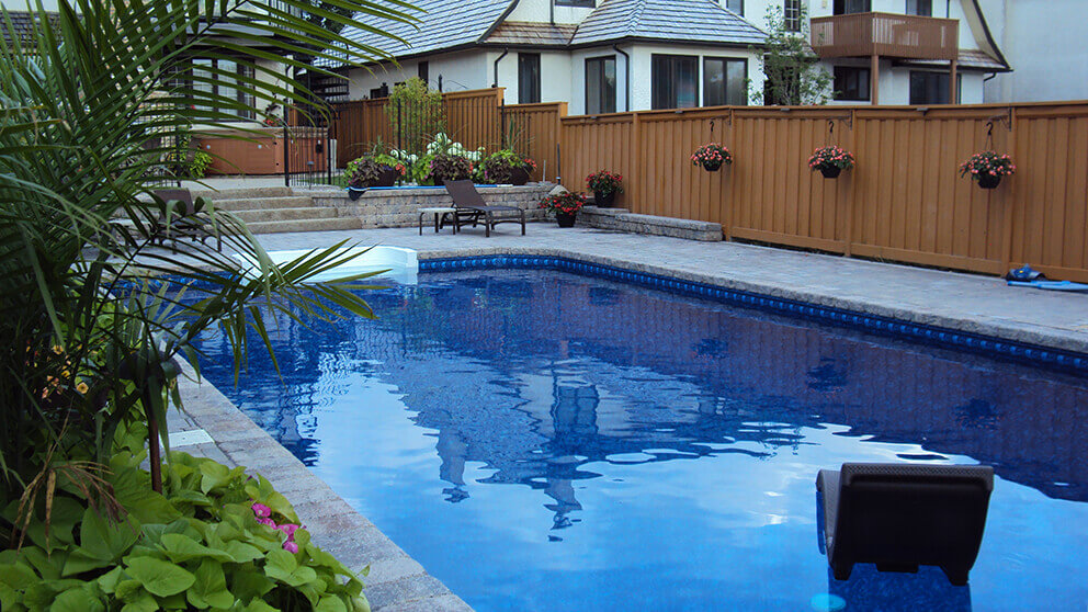 This bright backyard photograph captures a perfectly maintained in ground swimming pool that has just undergone a professional Winnipeg pool openings & closings service from Crown Spas and Pools. The still blue water reflects the afternoon sky and the large two storey white house in the background, while a black floating chair drifts in the lower right area. The pool is finished with a dark blue mosaic waterline tile and is surrounded by a vast deck of grey interlocking stone pavers. Lush greenery, including large palm fronds and vibrant pink flowers, fills the bottom left corner, adding a tropical texture to the scene. A light brown wooden fence encloses the yard, featuring several black hanging pots with red blossoms that contrast against the wood grain. The lighting is natural and even, highlighting the clean lines of the stone masonry and the pristine condition of the water. This residential setting is peaceful and serves as a prime example of expert seasonal pool maintenance in a suburban environment.