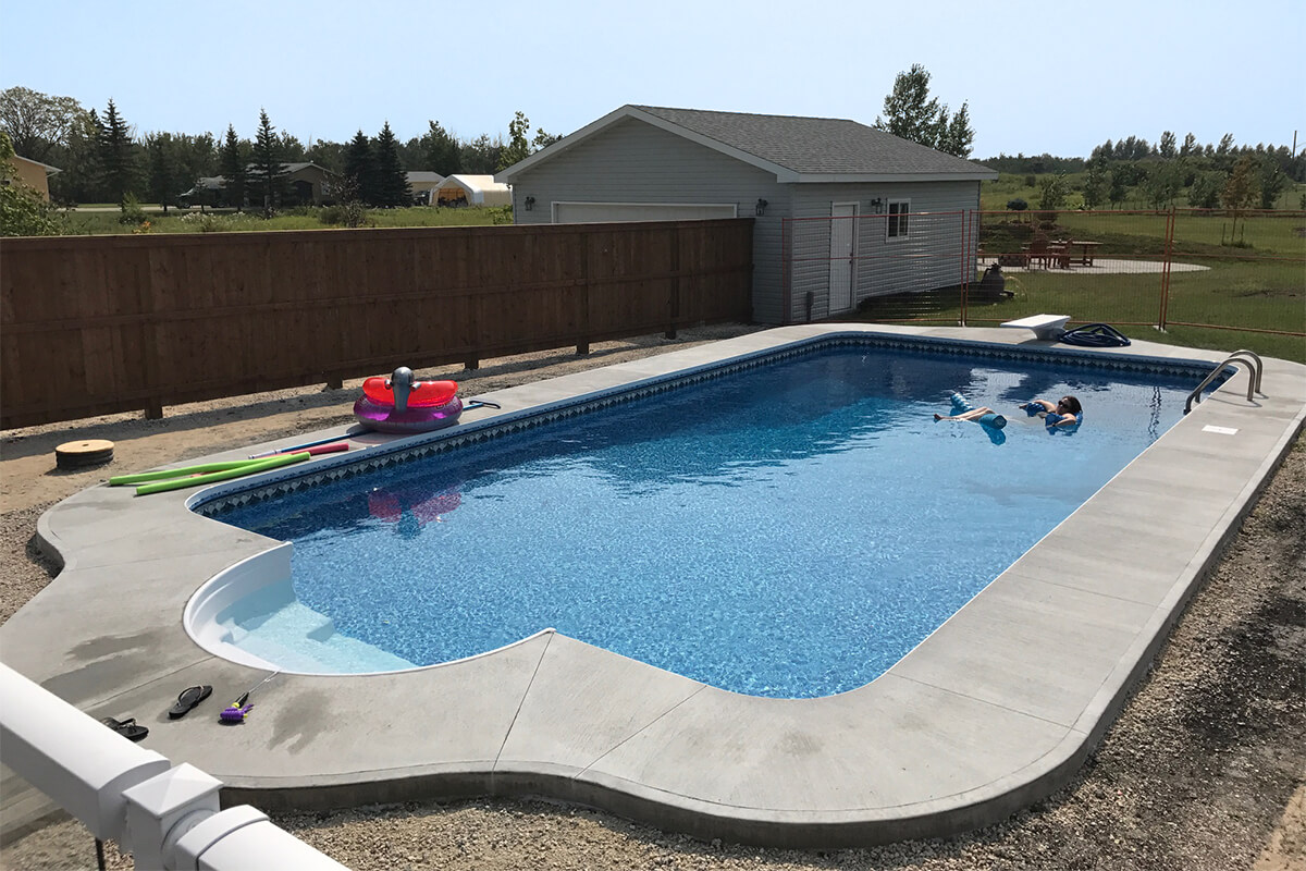 This outdoor wide angle photograph captures a sprawling residential backyard showcasing the newly installed Budd custom pool with Roman steps. The rectangular inground swimming pool is filled with rippling bright blue water and features a decorative blue patterned vinyl liner border along the waterline. At the near left end, a sweeping molded white entry area forms the Roman steps, which are seamlessly framed by a clean and continuous light grey concrete deck that completely surrounds the water. On the concrete deck near the steps in the bottom left corner, a pair of black flip flops and a small purple and green water toy rest next to a partial white railing. Further along the left edge of the pool, green and pink foam noodles and a bright pink and purple inflatable float are sitting on the concrete next to a long solid brown wooden fence. In the centre of the pool, a person wearing sunglasses floats comfortably on their back using a blue foam noodle in the deep water zone. The far right side of the concrete deck features a traditional white diving board, a coiled blue pool hose, and a stainless steel ladder with two curved handrails leading into the water. A white square skimmer lid is flush mounted into the concrete deck on the right edge. The immediate ground outside the concrete perimeter consists of unfinished rough gravel and dirt. The background environment consists of a light grey detached garage with a dark grey shingled roof and a white side door, an orange mesh construction fence tracing the right property line, and a distant tree line of coniferous and deciduous trees under a clear pale blue daytime sky.