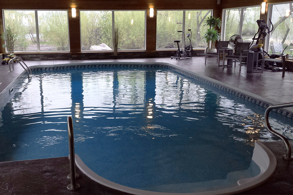 This eye-level perspective highlights the expansive layout of the Dacotah custom indoor pool with hot tub, emphasizing the versatility of the dedicated fitness and cardio space. The large, rectangular pool features a light-coloured moulded entry section in the bottom foreground and a stainless steel ladder on the left, set into a dark, textured floor. The right side of the deck functions as a personal fitness area, equipped with a stationary exercise bike, a pair of chairs, and an elliptical machine situated near the large windows. The room is defined by its warm wood-clad walls and floor-to-ceiling windows that provide natural light, creating an inviting atmosphere throughout the entire aquatic retreat. Artificial lighting is mounted along the wooden walls, complementing the outdoor brightness. Decorative potted plants and a small toy vehicle near the far left ladder add personal touches to this climate-controlled environment.