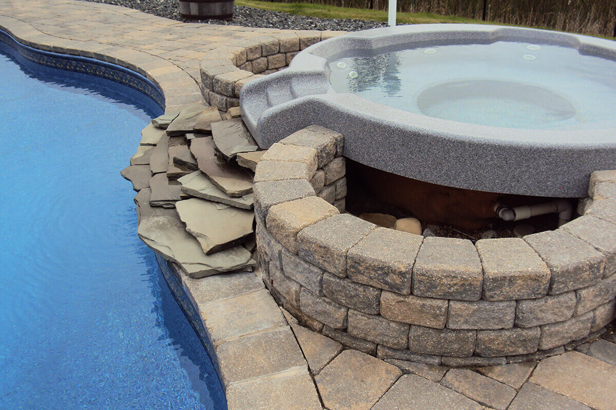 This close up photograph provides a detailed view of the elevated hot tub component of the Doral custom freeform pool with spa project. The round grey hot tub sits above the main pool level and is partially enclosed by a custom stone retaining wall constructed from textured tan and grey blocks. The spa features a smooth white interior with several circular jets positioned along the submerged inner wall, while no exterior panels or top side controls are visible from this angle. An opening in the stonework reveals the brown underside structural elements of the spa and its white PVC plumbing pipes in the shadows. A moulded grey spillway on the edge of the hot tub directs water over a series of strategically stacked flat natural rock slabs that bridge the gap down to the bright blue pool water on the left. The pool features a patterned blue mosaic tile border along the waterline. A massive interlocking block patio surrounds the installation, with a dark decorative rock bed and a blurred grassy background environment visible at the top of the frame.