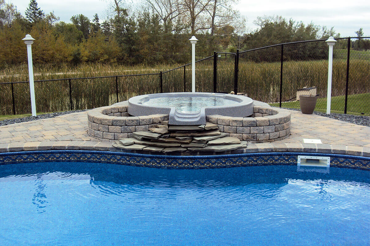 This eye level photograph perfectly frames the elevated hot tub of the Doral custom freeform pool with spa, looking directly across the bright blue swimming pool water. The grey hot tub acts as a central focal point on the large interlocking block patio, featuring a smooth interior with submerged circular jets spaced around the seating area. It is surrounded by a symmetrical custom stone retaining wall, and water is designed to flow from the front hot tub spillway over a cascading waterfall made of flat stacked natural rock slabs directly into the pool below. A white rectangular skimmer is built into the pool wall on the lower right, positioned just below the blue decorative tile border. Dark decorative rock beds flank the left and right sides of the stone patio. In the background environment, a sleek black safety fence runs horizontally across the yard, accented by three tall white light posts, separating the pristine outdoor living space from a natural wetland area filled with tall marsh grasses and bare trees under an overcast grey sky.