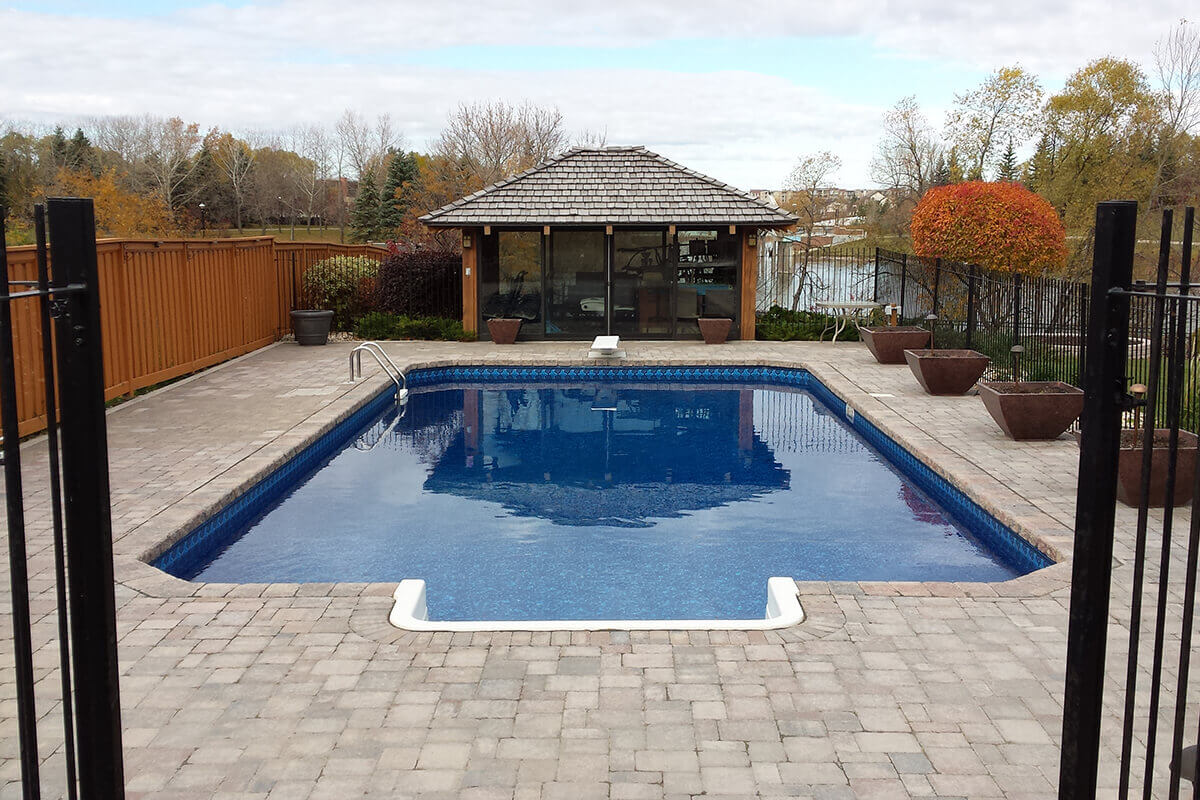This straight on outdoor photograph looks down the length of the magnificent Shorecrest inground pool with paving stones, framed by the open gates of a black metal safety fence in the foreground. At the bottom centre, the crisp white walk-in step section is built into the near edge of the clear blue pool water. The massive, beautifully patterned stone paver deck borders the entire pool, expanding outwards on both sides. A solid brown wooden fence runs along the left property line, while large square brown planter bowls decorate the right side of the deck near another black metal fence. At the far end of the pool in the centre background, a classic white diving board sits on the deck just in front of an incredible glass-walled pool house featuring a brown wooden shingle roof. Through the transparent pool house and the surrounding bare autumn trees, a calm lake is visible under a bright, partly cloudy sky.