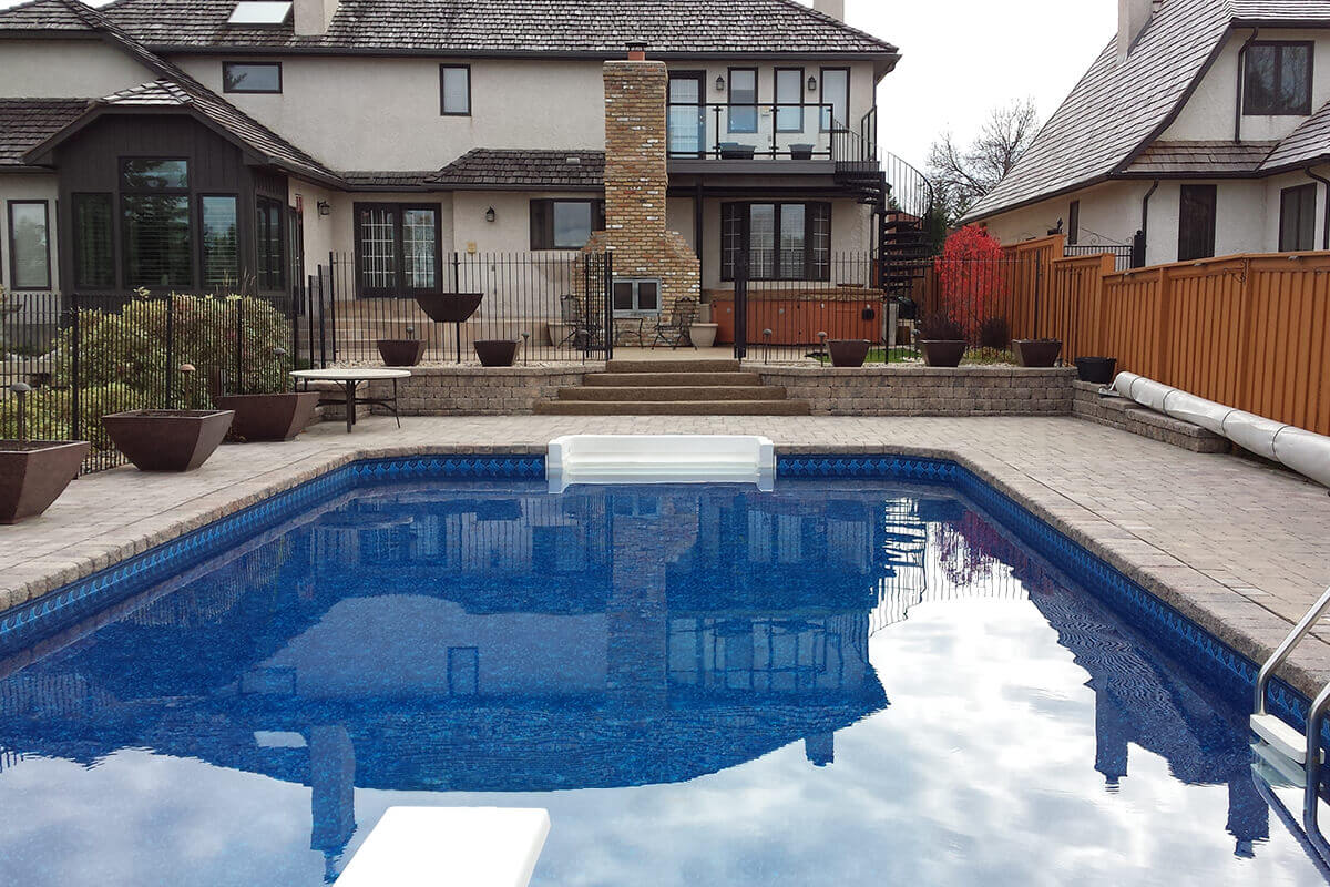 This outdoor photograph looks across the clear blue water of the Shorecrest inground pool with paving stones towards the tiered luxury estate home. In the bottom left corner, the tip of a classic white diving board extends over the deep end of the water. The massive patterned stone paver deck wraps around the pool, featuring a crisp white walk-in step section situated perfectly in the middle of the far pool edge. Just beyond the pool deck, a low stone retaining wall and a set of wide concrete steps lead up through a black metal safety fence to an expansive upper terrace tier. On this elevated patio, a prominent tall brick outdoor fireplace stands in the centre, flanked by a premium wood skirted hot tub on the right and an outdoor patio table on the left. The large beige stucco house with dark brown trim and a wood shingle roof forms the entire background, complete with a spiral metal staircase on the right leading to a second story balcony.