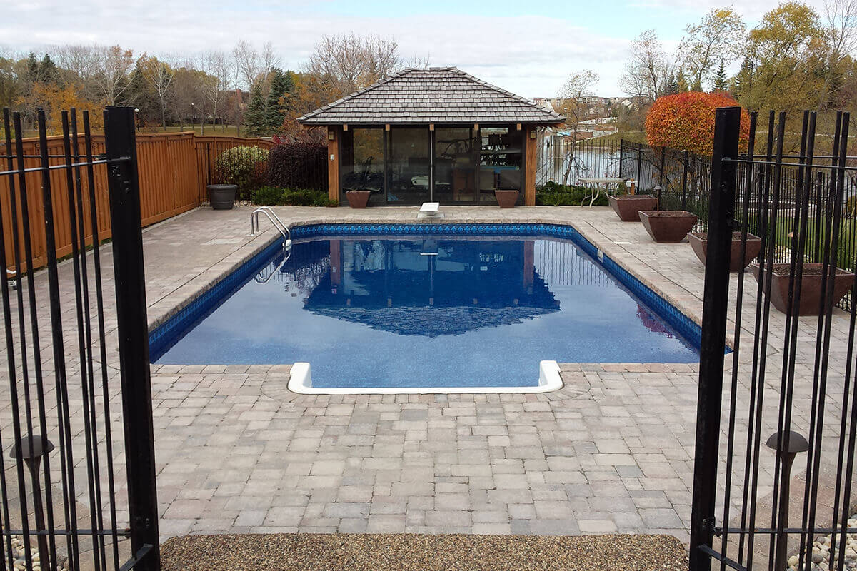 This symmetrical outdoor photograph provides a beautifully centred view down the length of the expansive Shorecrest inground pool with paving stones. The shot is framed perfectly between two tall black metal fence posts in the immediate left and right foreground. A massive, beautifully patterned stone paver deck surrounds the bright blue water, beginning with the crisp white walk-in step section at the near edge. The pool extends towards the centre background, where a classic white diving board is mounted on the deck. Directly behind the diving board sits the incredible glass-walled pool house with a brown wood shingle roof, offering a sheltered entertainment space. The left side of the pool area is bordered by a tall, solid brown wooden fence, while the right side features decorative brown square planters lining a black metal fence. In the distant background, a tranquil lake and autumn trees are visible under a bright, partly cloudy sky.