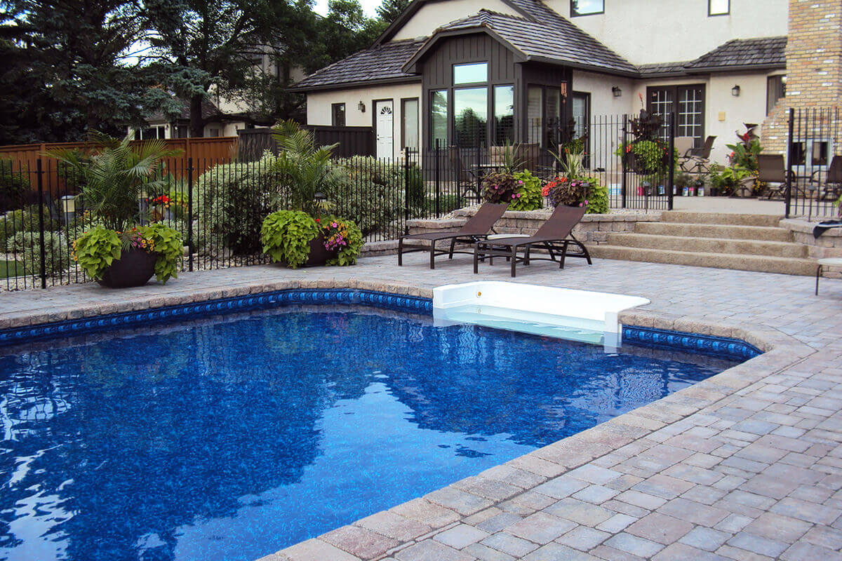 This angled outdoor photograph highlights the relaxing lounging area beside the Shorecrest inground pool with paving stones. The bright blue swimming pool fills the foreground and left side, with a crisp white walk-in step section integrated into the right edge for effortless entry. A massive patterned stone paver deck borders the water, featuring two dark brown modern lounge chairs sitting side by side on the deck just past the pool steps. Several large, dark planters overflowing with vibrant green foliage and pink flowers are strategically placed along the deck near a black metal safety fence. Behind the safety fence, wide concrete steps lead up to a tiered patio level. On the upper right, a portion of the large beige stucco house is visible, showcasing the tall brick outdoor fireplace. A solid brown wooden fence and mature green trees enclose the left side of the sprawling backyard resort.