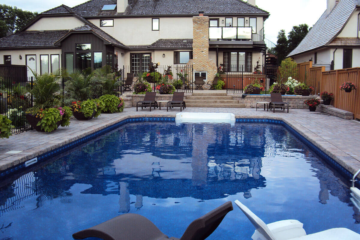 This eye level outdoor photograph looks across the rippling blue water of the Shorecrest inground pool with paving stones towards the multi-level entertainment space. In the immediate foreground, the tops of two dark brown floating pool lounge chairs are visible in the deep water. The pool is surrounded by a massive, beautifully patterned stone paver deck, with a crisp white walk-in step section centred on the opposite side. Beyond the pool, a low stone retaining wall and wide stairs lead up to an expansive upper terrace tier, separated by a black metal safety fence adorned with lush potted plants. This upper patio features multiple dark brown outdoor lounge chairs, a premium hot tub on the right, and a towering brick outdoor fireplace in the centre attached to the large beige stucco house. A solid brown wooden fence runs along the right property line under a bright, partly cloudy sky.