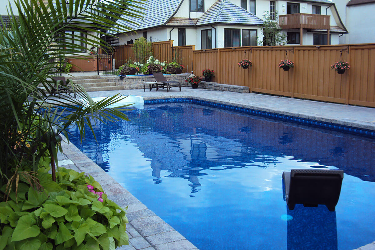 This beautiful outdoor photograph captures a vibrant section of the Shorecrest inground pool with paving stones. In the bottom left corner, a large dark planter bursts with lush green tropical plants and pink flowers, sitting on the massive patterned stone paver deck. The bright blue water of the pool occupies the majority of the frame, featuring a dark brown floating lounge chair resting on the surface in the bottom right area. On the far side of the pool, the deck includes a crisp white walk-in step section leading into the water. Behind the pool deck, a tall, solid brown wooden fence stretches across the right background, decorated with several hanging baskets filled with pink flowers. On the left side, stone steps lead up to a tiered patio area where a dark lounge chair sits. The large beige stucco estate home with a wooden balcony is visible in the upper background behind the wooden fence.