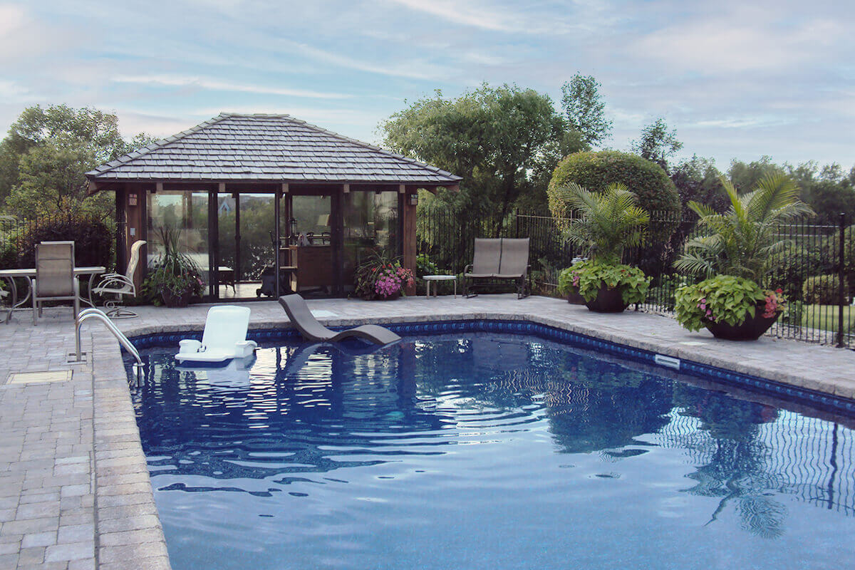 This eye level outdoor photograph showcases the relaxing atmosphere of the Shorecrest inground pool with paving stones. The clear blue water of the swimming pool fills the foreground, featuring a crisp white floating pool chair on the left and a dark brown floating lounge chair on the right. A massive, beautifully patterned stone paver deck surrounds the water, complemented by a stainless steel pool ladder on the left edge. At the far end of the pool deck, an incredible glass-walled pool house with a dark brown wood shingle roof provides a shaded retreat, furnished with a patio dining set on the left and two seating chairs on the right. Lush tropical potted plants with bright green leaves and pink flowers decorate the deck space near a black metal safety fence on the right. In the background, a tranquil lake surrounded by dense green trees creates a serene, private resort environment under a soft cloudy sky.
