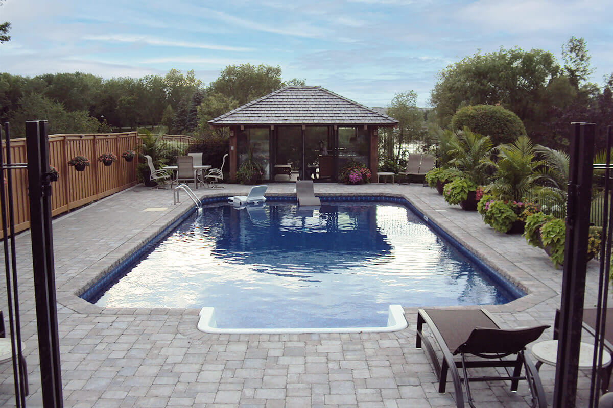 This wide angle outdoor photograph provides a comprehensive view down the length of the Shorecrest inground pool with paving stones. The shot is framed by tall black metal fence posts on the near left and right edges. In the bottom right foreground, a dark brown outdoor lounge chair sits on the massive, beautifully patterned stone paver deck that completely surrounds the water. The bright blue pool features a crisp white walk-in step section at the near bottom centre, while a white and a dark brown floating lounge chair drift comfortably in the middle of the water. At the far end of the pool, the incredible glass-walled pool house with a wood shingle roof is flanked by large tropical potted plants. A solid brown wooden fence runs down the left side of the yard, and a black metal safety fence runs down the right. Mature green trees and a calm lake complete the distant background under a cloudy daytime sky.